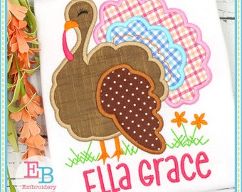 Puede incluir: Una camiseta blanca con un diseño de pavo bordado. El pavo tiene plumas marrones, con lunares y a cuadros, con patas naranjas y una carúncula rosa. El nombre "Ella Grace" está bordado en rosa debajo del pavo, con hierba verde y flores naranjas.