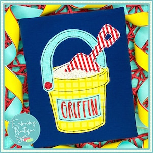 May include: A yellow and blue embroidered bucket and shovel with the name "Griffin" on the front. The bucket and shovel are on a dark blue background.