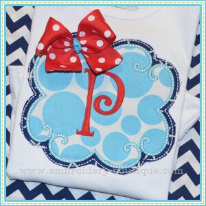 Puede incluir: Una camisa blanca con un diseño de lunares azules y blancos y un lazo rojo. La camisa tiene un diseño bordado azul con la letra "P" en el centro. El texto "www.embroideryboutique.com" está impreso en la camisa.