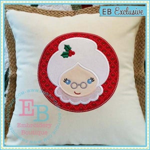May include: White pillow with a red and white embroidered circle featuring a cartoon image of a woman with white hair, glasses, and a red holly sprig. The text "EB Exclusive" is in the upper right corner of the pillow.