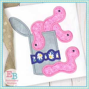 May include: Embroidered design of a pink worm spilling out of a gray can with a blue and white bow. The worm has a swirl pattern.
