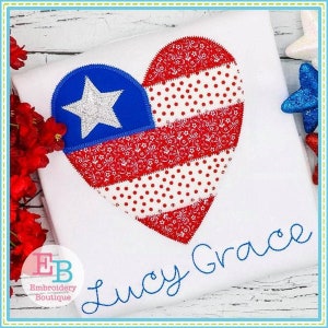 May include: A white t-shirt with a red, white, and blue heart-shaped American flag design. The heart is made up of different patterned fabrics, including polka dots and floral prints. The name "Lucy Grace" is embroidered in blue cursive script below the heart.