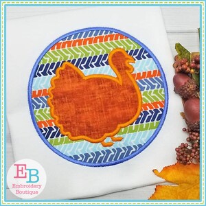 May include: Embroidered design of a brown turkey in a blue, green, and orange circle with a chevron pattern. The design is on a white fabric.