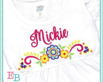 May include: White sleeveless shirt with a pink embroidered name "Mickie" and a colorful floral design with swirls.