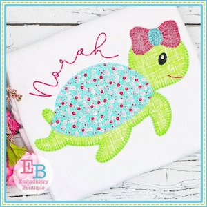 May include: A green and blue applique of a turtle with a pink bow on its head. The turtle is on a white background with the name "Norah" embroidered in pink.