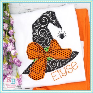 Puede incluir: Un sombrero de bruja negro y blanco con un lazo verde y naranja de lunares y una araña en la parte superior. El nombre "Elyse" está bordado debajo del sombrero.