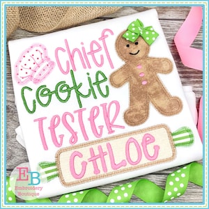 May include: A white t-shirt with pink and green embroidery that reads "chief cookie tester CHLOE". The design includes a gingerbread person with a green bow and a pink and white chef's hat.