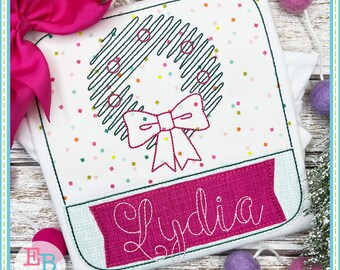 Op de afbeelding: Een wit shirt met een groene en zwarte krans met een roze strik en een roze banner met de naam "Lydia" geborduurd.
