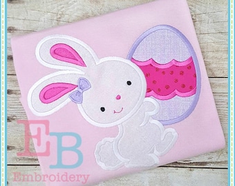 May include: A white bunny with pink ears and a pink bow holding a purple and pink Easter egg with white polka dots. The bunny and egg are embroidered on a pink shirt.