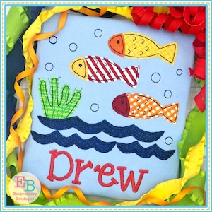 Peut inclure: Un tissu bleu avec un nom personnalisé "Drew" brodé en rouge. Le motif représente trois poissons orange et rouge nageant dans une eau bleue avec des algues vertes.