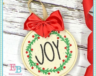 May include: A round, cream-coloured ornament with a green and red wreath and the word "JOY" embroidered in black. The ornament is hanging from a red ribbon with a bow.