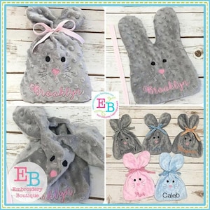 May include: A set of gray bunny-shaped plush bags with embroidered names. The bags are decorated with pink and blue bows and have a pink nose and black eyes. The names embroidered on the bags are Brooklyn, Casey, Levi, Ava, and Caleb.
