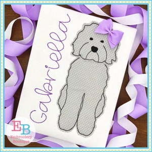 May include: Embroidered design of a gray dog with a purple bow on a white t-shirt. The name "Gabriella" is embroidered in purple on the shirt.