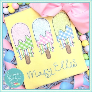 May include: A yellow t-shirt with three embroidered popsicles with bows. The popsicles are pink, blue, and green. The text "Mary Ellis" is embroidered below the popsicles.