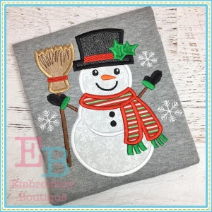 May include: A white snowman with a black top hat, red and green striped scarf, and a broom. The snowman is smiling and has two black buttons. The background is a gray fabric with snowflakes.