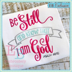 May include: Embroidered design on a white t-shirt with the text "Be Still and Know That I am God" in pink and teal lettering with a gray banner and the text "Psalm 46:10" in black.