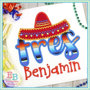 May include: White t-shirt with a colorful embroidered design. The design features a red sombrero above the word "tres" in a striped pattern, with the name "Benjamin" below in red. The design is outlined in blue.