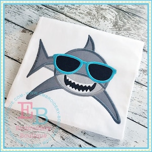 May include: A gray shark with a toothy grin wearing blue sunglasses. The shark is embroidered on a white t-shirt.