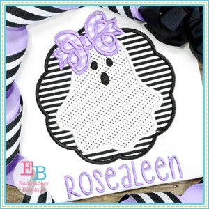 May include: A white ghost with a purple bow on its head. The ghost is outlined in black and has a black face with two eyes and a mouth. The ghost is on a white and black striped background. The name "Rosealeen" is embroidered in purple below the ghost.