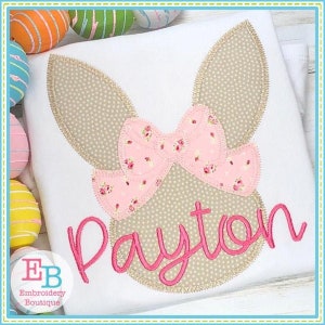 Pode incluir: Camiseta branca com um design de rosto de coelho. Apresenta orelhas bege, um laço floral rosa e o nome "Payton" bordado em rosa. O design é rodeado por uma costura dourada.