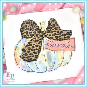 May include: A blue and yellow watercolor pumpkin with a gold glitter design and a brown leopard print bow. The pumpkin has a tag that says "Sarah".