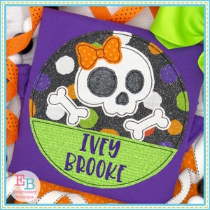 May include: A purple shirt with a round applique featuring a white skull with an orange bow, green and orange polka dots, and two white bones. The applique has a green banner with the text "IVEY BROOKE" in white.