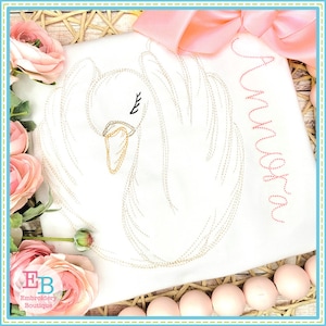 May include: White embroidered swan design on a white fabric with the name "Aurora" embroidered in pink thread.
