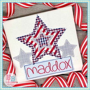 May include: A white t-shirt with a red, white, and blue star applique. The star is layered with a red and white striped fabric, a blue and white gingham fabric, and a gray fabric. There are two smaller gray stars on either side of the large star. Below the stars is a blue and white striped banner with the name "maddox" embroidered in red.