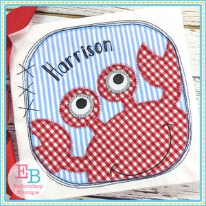May include: Embroidered applique of a red and white gingham crab with a smiling face and large eyes. The crab is on a blue and white striped background with the name "Harrison" embroidered above it.