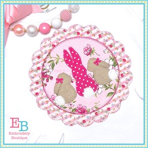 Pode incluir: Uma aplicação de tecido rosa e branco com dois coelhos. Os coelhos estão rodeados de flores cor-de-rosa e folhas verdes. A aplicação está sobre um fundo branco.