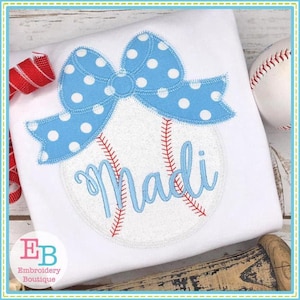 Puede incluir: Un balón de béisbol blanco con costuras rojas y un lazo azul con lunares blancos. El nombre "Madi" está bordado en el balón de béisbol con hilo azul.