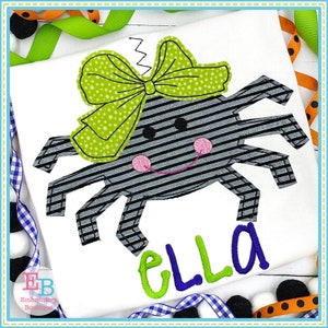 May include: A black and white striped spider with a green bow and a pink smiling face. The spider has eight legs and is wearing a bow on its head. The word "ELLA" is embroidered in green and blue below the spider.