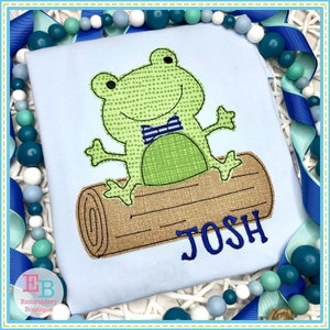 May include: A light blue t-shirt with an embroidered green frog wearing a blue bow tie sitting on a brown log. The name "JOSH" is embroidered in blue below the frog.