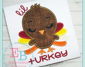 Puede incluir: Tela blanca con un diseño bordado de un pavo de dibujos animados. El pavo es marrón con un patrón texturizado, un pico amarillo y plumas de cola coloridas. Las palabras "lil" y "+ turkey" están bordadas en rojo.
