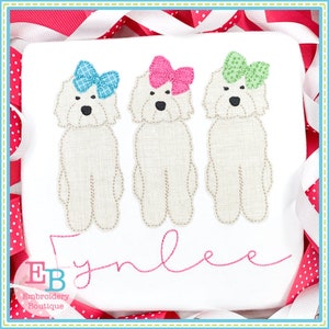May include: Three white dogs with bows on their heads. The dogs are standing in a row. The bows are blue, pink, and green. The name "Emilee" is embroidered below the dogs.