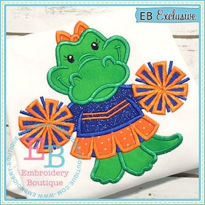 May include: Embroidered design of a green alligator wearing a blue and orange cheerleader uniform and holding orange and blue pom-poms. The design includes the text "EB Exclusive".