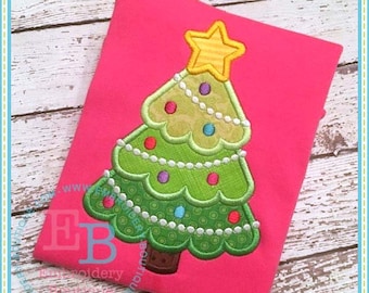 May include: A green Christmas tree with a yellow star on top. The tree is decorated with white beads, red, green, and blue ornaments. The tree is embroidered on a pink shirt.