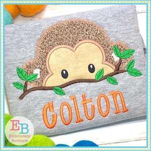May include: Embroidered design of a brown and tan hedgehog peeking out from behind a branch with green leaves. The name "Colton" is embroidered in orange below the hedgehog.