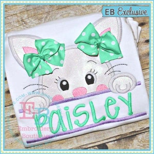 May include: White bunny peeking over a banner with the name "Paisley" embroidered in green. The bunny has green and white polka dot bows on its ears and pink cheeks.