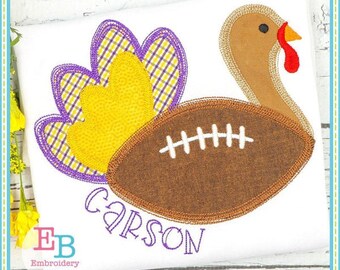May include: A white t-shirt with a turkey design incorporating a football. The turkey has a brown head, a red wattle, and a tail with purple and yellow detailing. The name "Carson" is embroidered in purple script below the football.