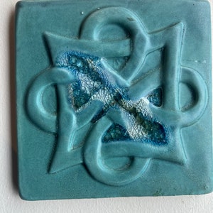 May include: A teal blue ceramic tile with a raised, intricate Celtic knot design. The center of the knot is filled with a crackled glaze in shades of blue and white.