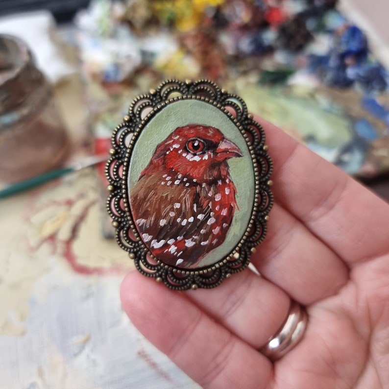 May include: A small, round, bronze-colored brooch with a detailed design. The brooch features a painted image of a red and brown bird with white spots.
