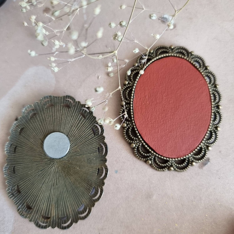 May include: Two ornate metal frames with a decorative design. One frame is empty and the other has a red backing.