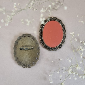 May include: Two antique-style brooches with ornate metal frames. One brooch has a red oval cameo, the other has a detailed, sunburst-like design.