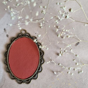 May include: A red oval cameo with a decorative bronze frame. The frame has a scalloped edge and a delicate floral design.