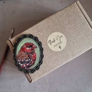 May include: A hand-painted bird brooch with red, white, and brown details. The bird is set in a bronze-colored oval frame with a decorative edge.