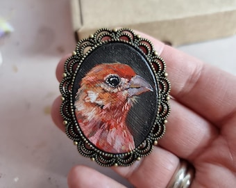 Op de afbeelding: Een handgeschilderde vogelbroche met een rode en oranje vink op een zwarte achtergrond. De broche is gezet in een ovaal bronzen frame met een decoratieve rand.