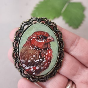 May include: A hand-painted bird brooch with a red and brown feathered design, set in a bronze oval frame with a decorative border.