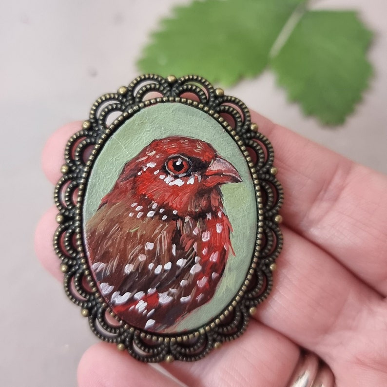 May include: A hand-painted bird brooch with a red and brown feathered design, set in a bronze oval frame with a decorative border.