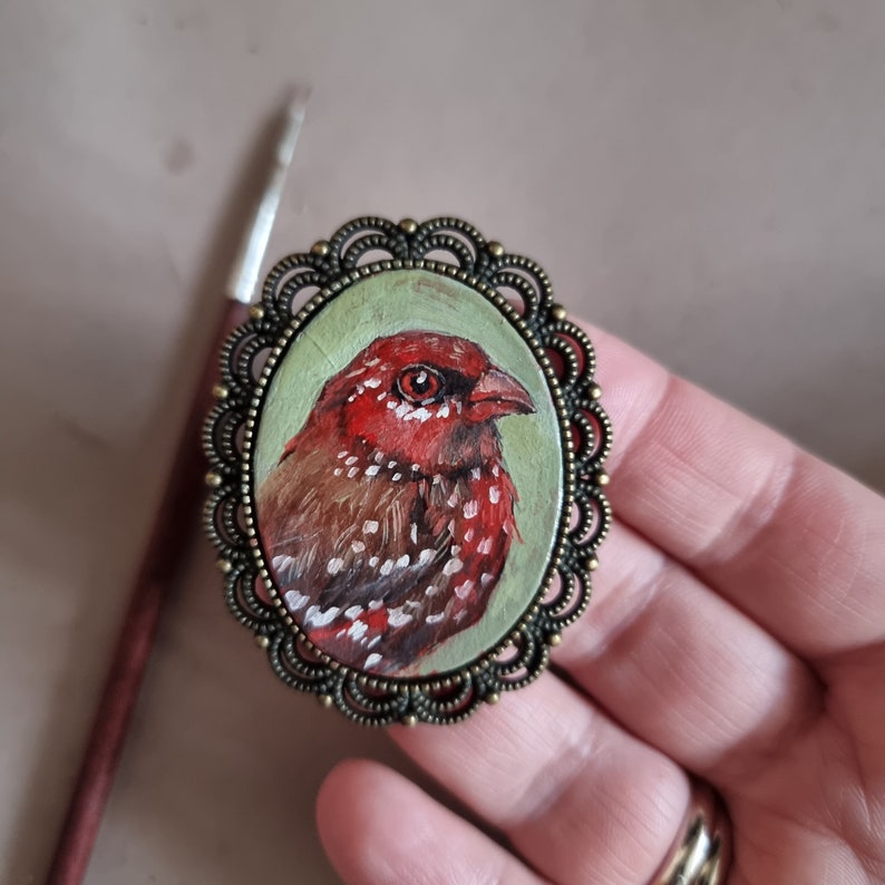 May include: A small, round, bronze-colored brooch with a painted image of a red and brown bird with white spots. The bird is set against a green background.